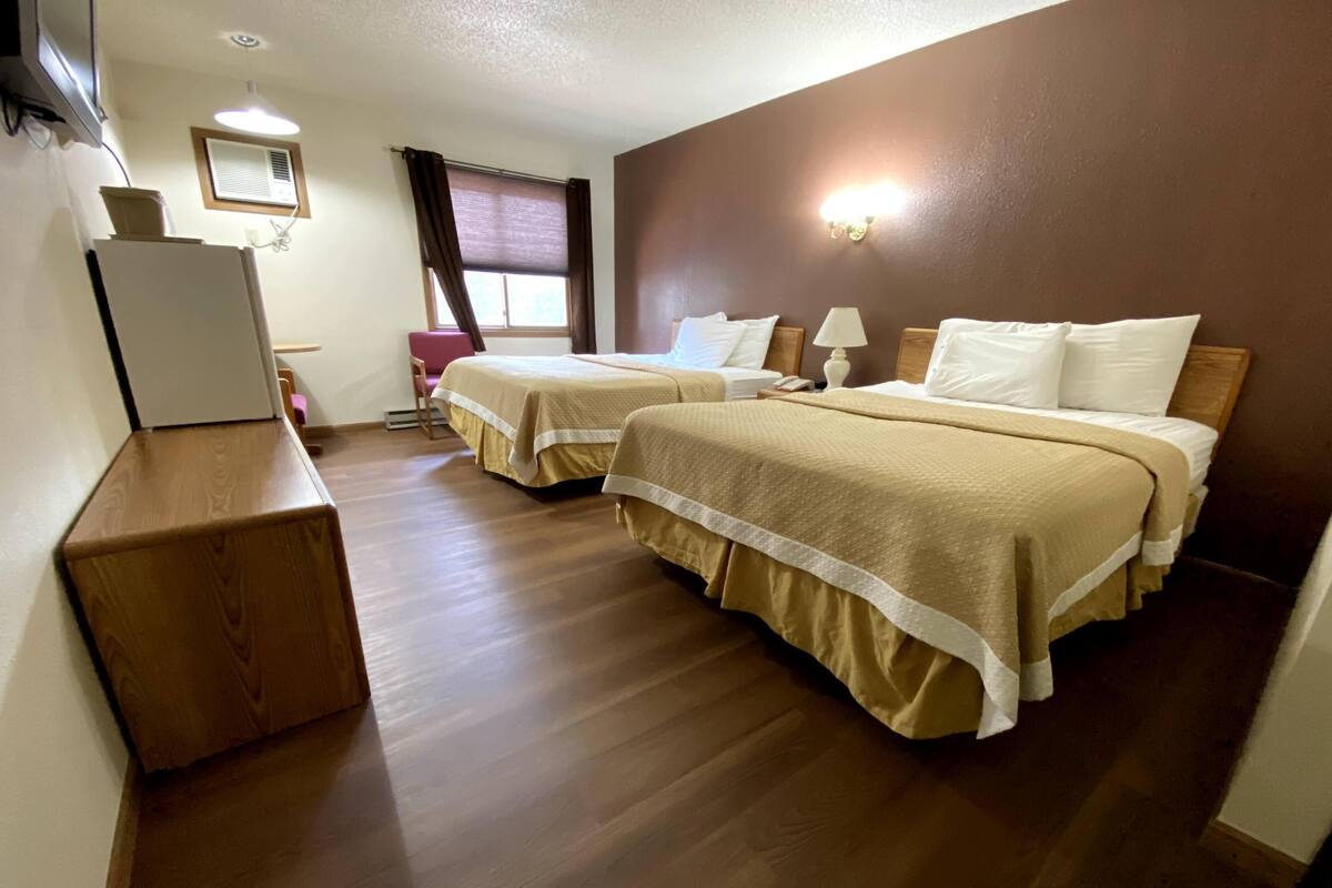 A hotel room with two beds, wooden flooring, a desk, a chair, a window with curtains, and a wall-mounted lamp.