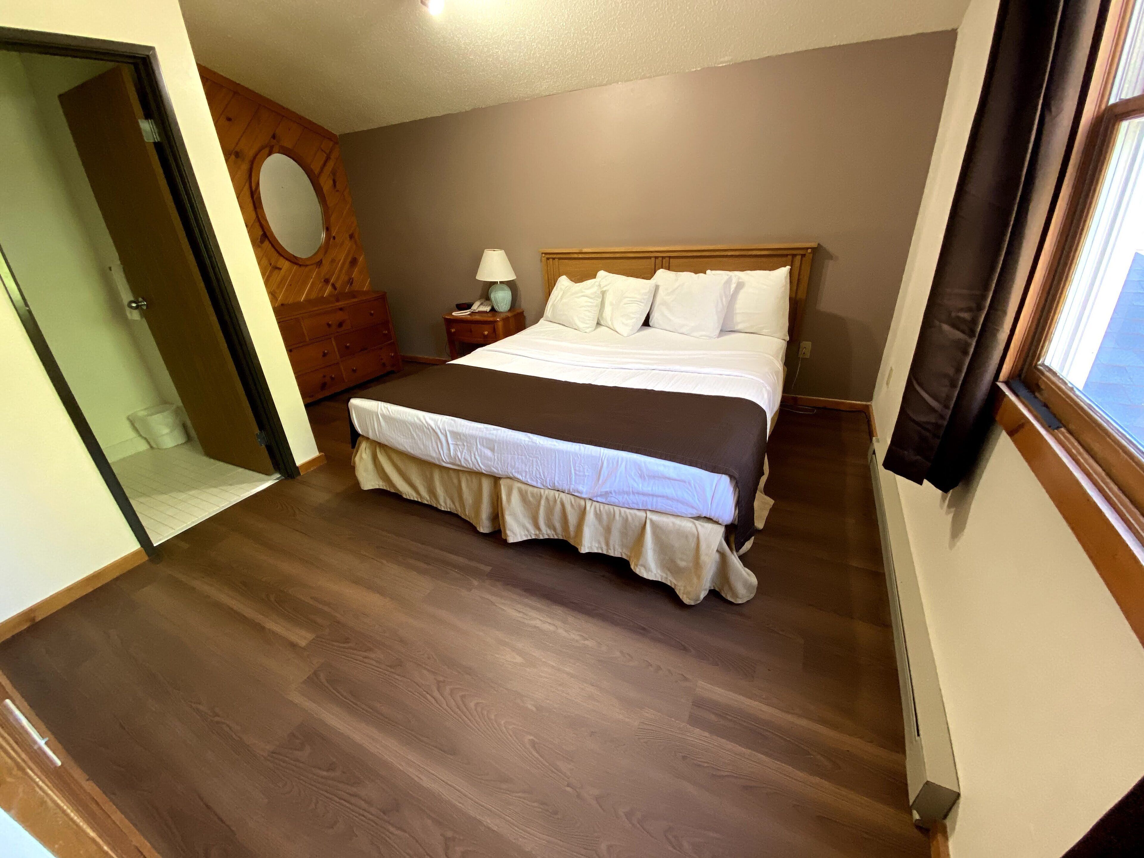 A bedroom with a large bed, wooden floor, a wooden dresser, a nightstand, a lamp, and a window with curtains.