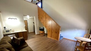 A cozy living room with a wooden staircase, a kitchen area with a microwave, and a dining table with a chair.
