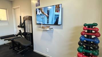 Un gimnasio en casa con equipo de ejercicio, un televisor montado en la pared y un estante con mancuernas.