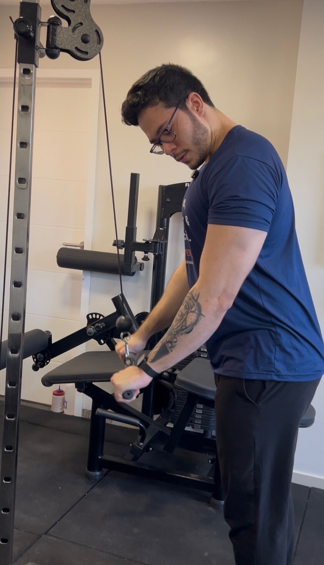 Eine Person trainiert mit einer Kabelzugmaschine in einem Fitnessstudio, im Hintergrund sind eine Tür und eine Wand zu sehen.