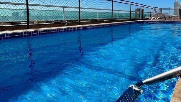 Una piscina azul cristalina con barandilla metálica, con vistas a un paisaje urbano con edificios y una masa de agua.