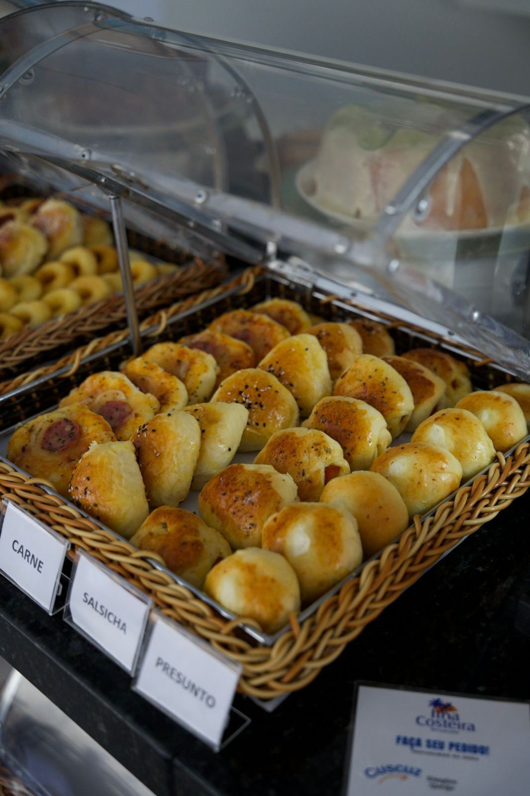Buffet com diversos pães e produtos de panificação, incluindo rolinhos rotulados 'CARNE', 'SALSICHA' e 'PRESUNTO'.