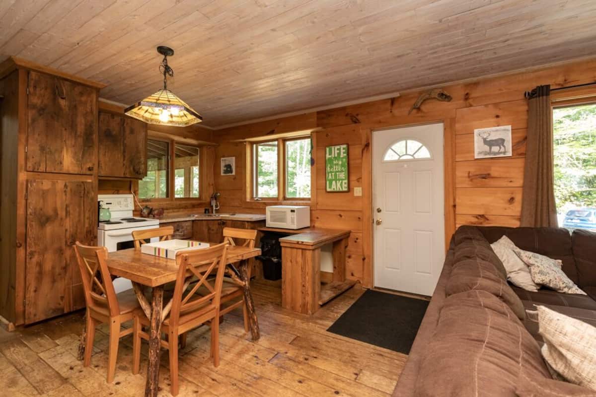 A cabin interior with a wooden dining table, chairs, a kitchen area, a sofa, and a door with a window.