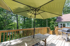 A wooden deck with a table and an umbrella, a grill, and a house in the background.