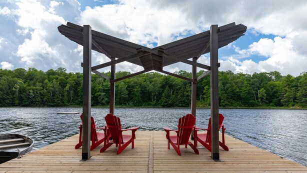 Une jetée en bois, agrémentée de deux fauteuils Adirondack rouges placés sous une pergola en bois, offre une vue sur un lac dont les rives sont bordées d’une forêt.