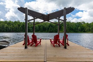 Une jetée en bois, agrémentée de deux fauteuils Adirondack rouges placés sous une pergola en bois, offre une vue sur un lac dont les rives sont bordées d’une forêt.