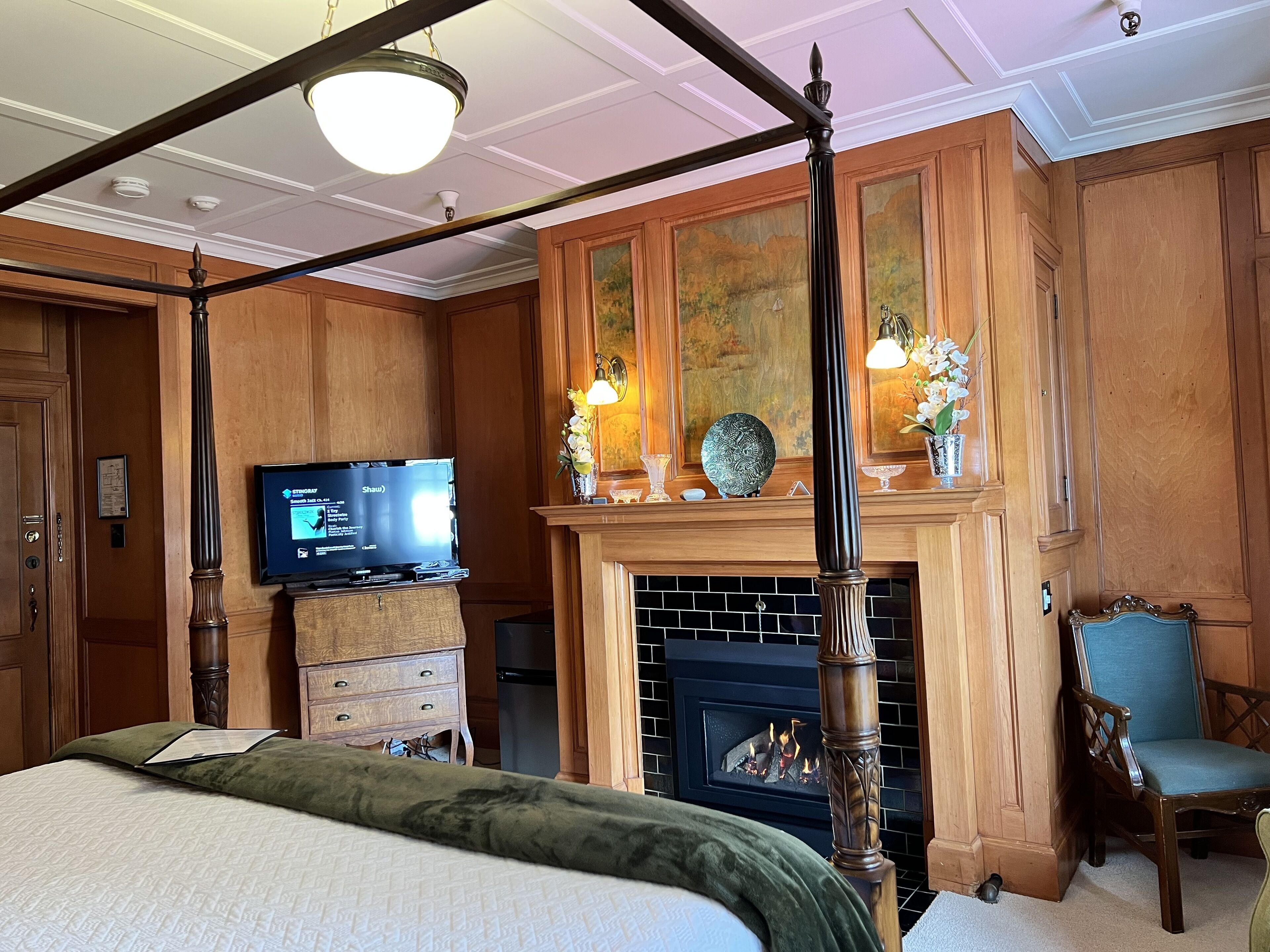 A four-poster bed, a television, a fireplace, and a chair in a room with wooden paneling and a ceiling fan.