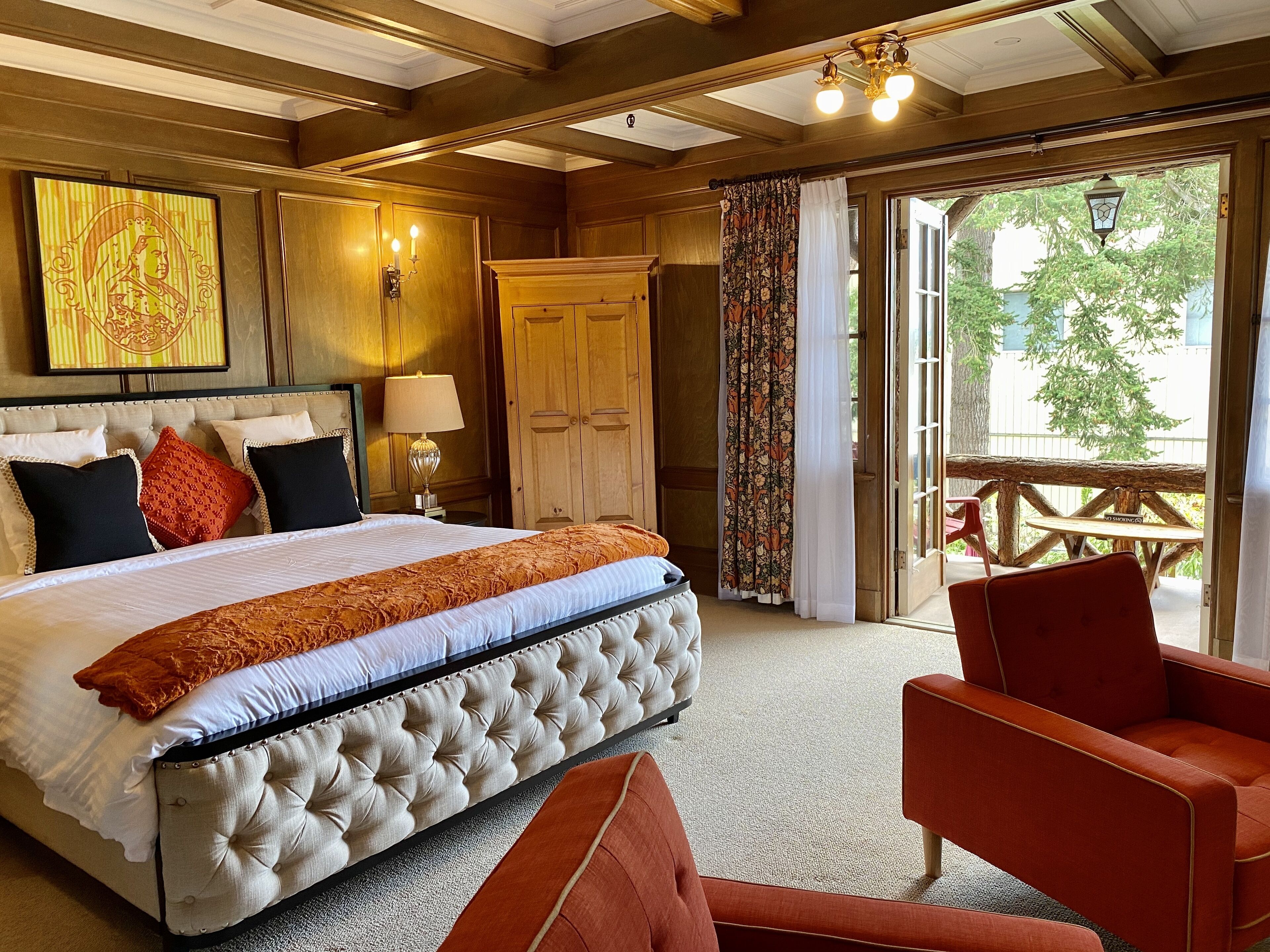 A hotel room with a large bed, a wooden wardrobe, a sitting area with a red chair, and a view of the outdoors through a glass door.