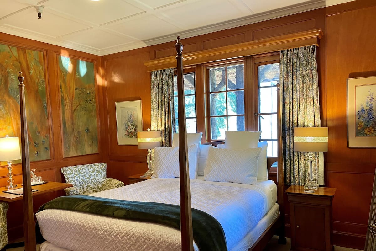 A four-poster bed with a quilted bedspread and multiple pillows, flanked by nightstands with lamps, in a room with wooden paneled walls and large windows with curtains.