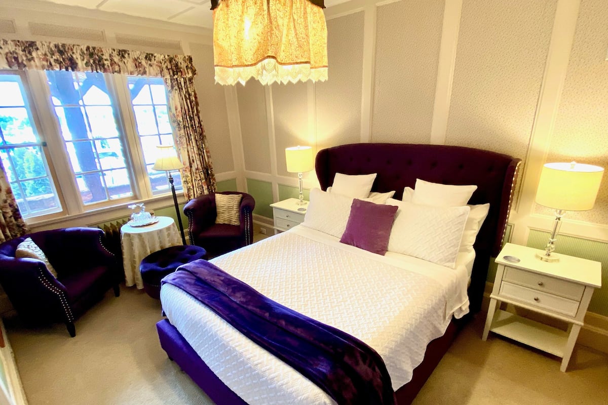 A well-lit bedroom with a large bed, two bedside tables, a chair, and a window with curtains.