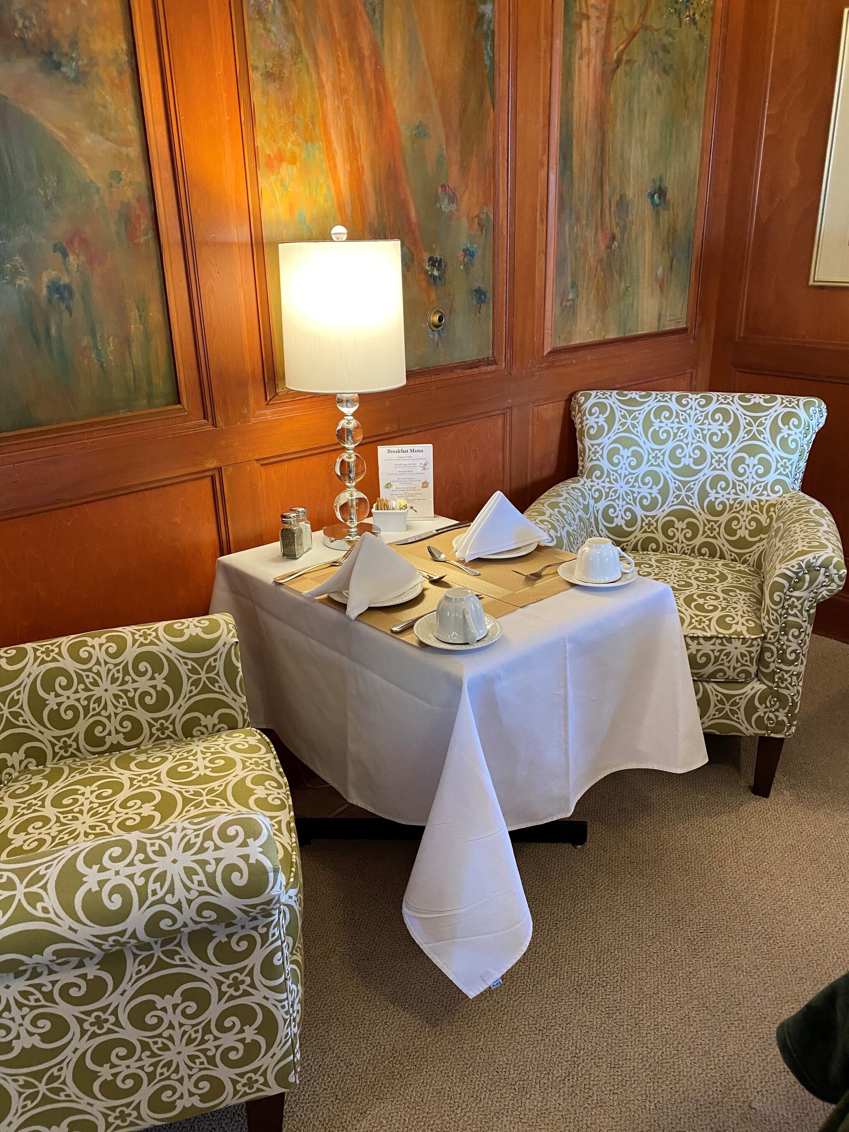 Un coin repas avec une table dressée pour deux personnes, une lampe et un menu de petit-déjeuner.