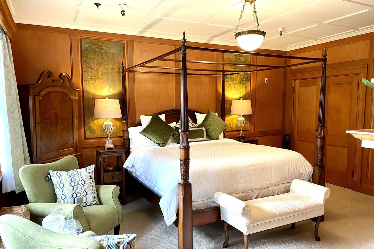 A four-poster bed with a canopy, two bedside tables with lamps, a green armchair, a white bench, and a wooden wardrobe.