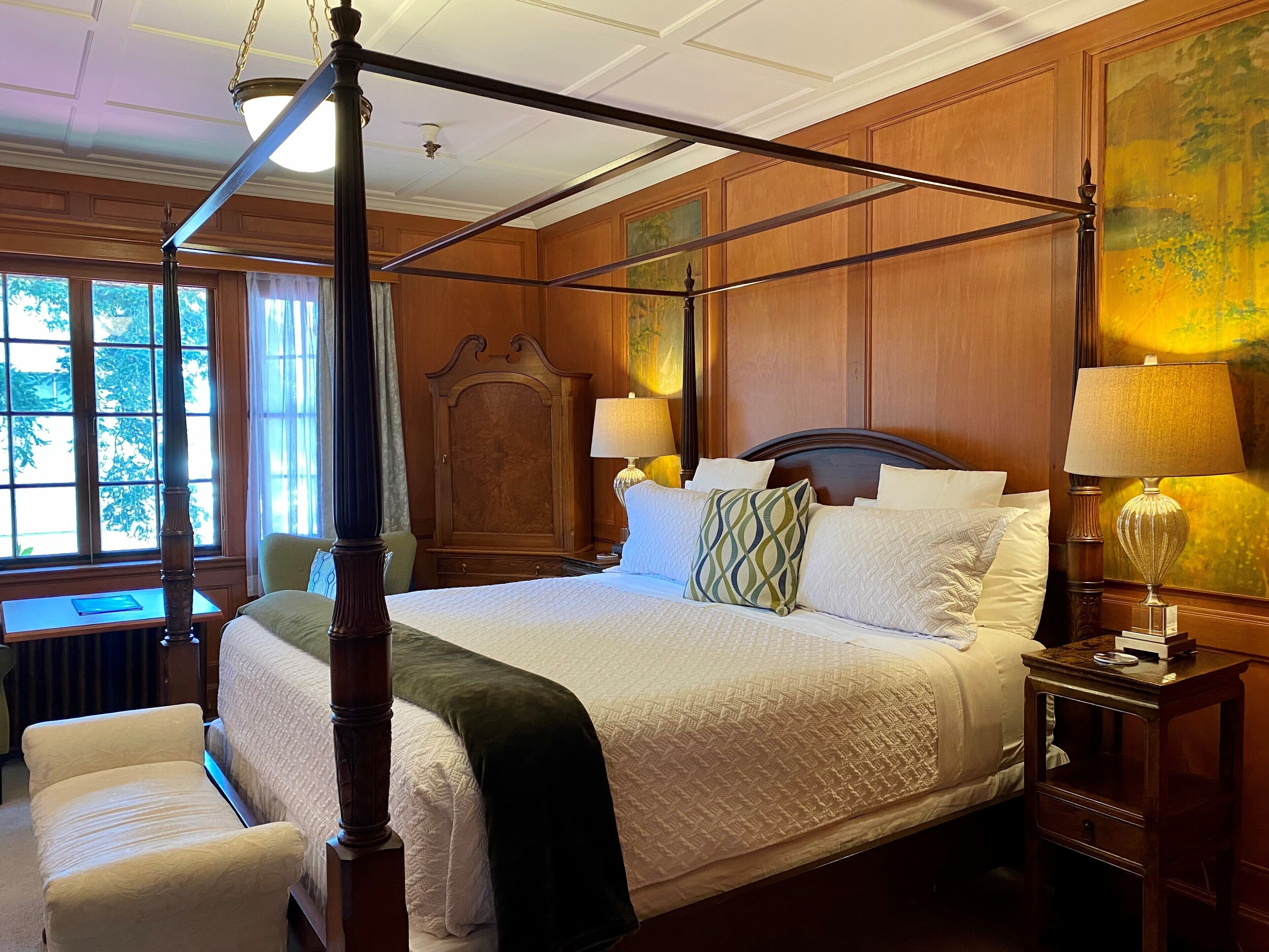A four-poster bed with a canopy, a wooden nightstand with a lamp, and a view of the outdoors through large windows.