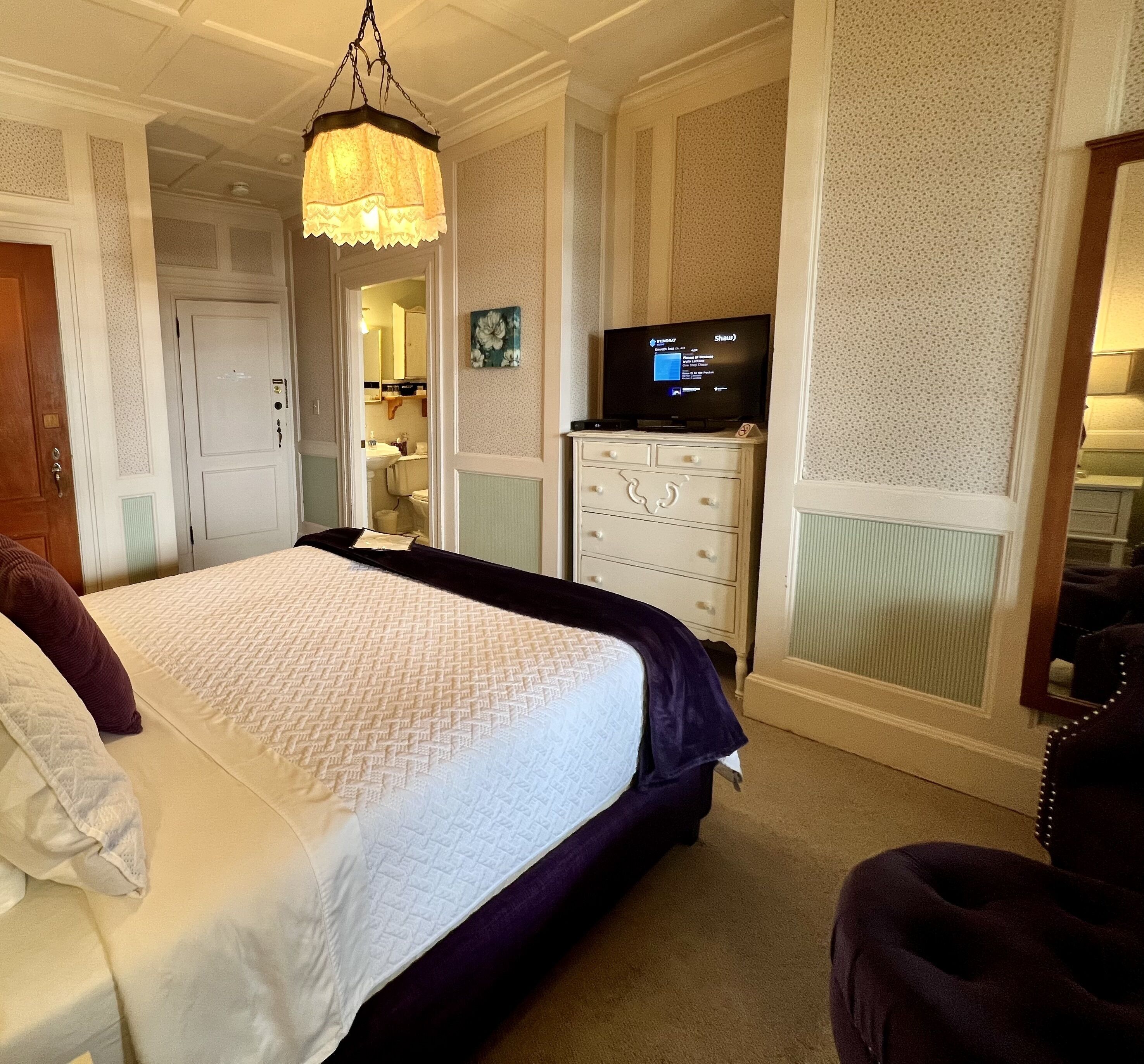 A bedroom with a large bed, a dresser, a television, and a bathroom visible through an open door.