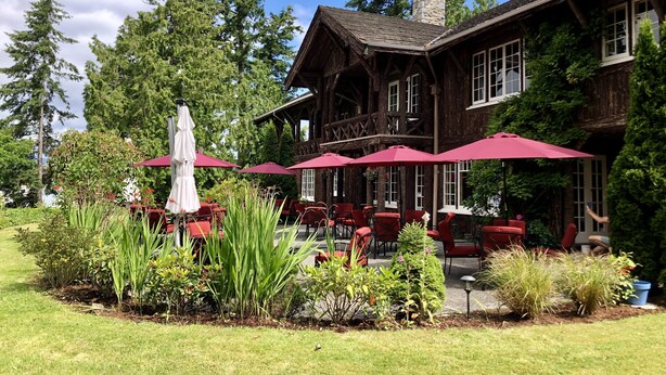 A traditional wooden building with a stone chimney, outdoor seating with red umbrellas, and lush greenery. - The Latch Inn (Sidney)