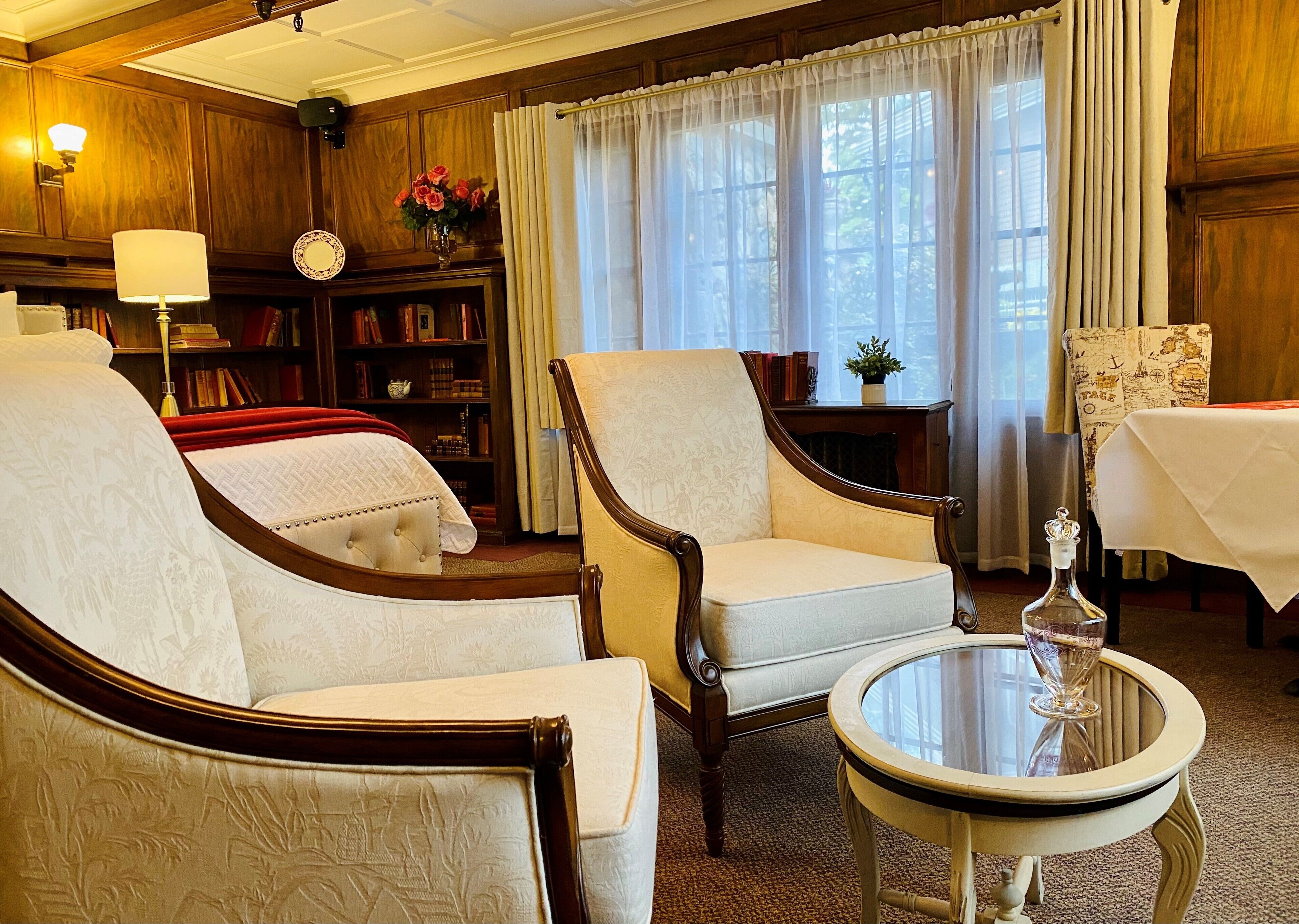 A traditional room with two armchairs, a round table, a bookshelf, and a window with curtains.