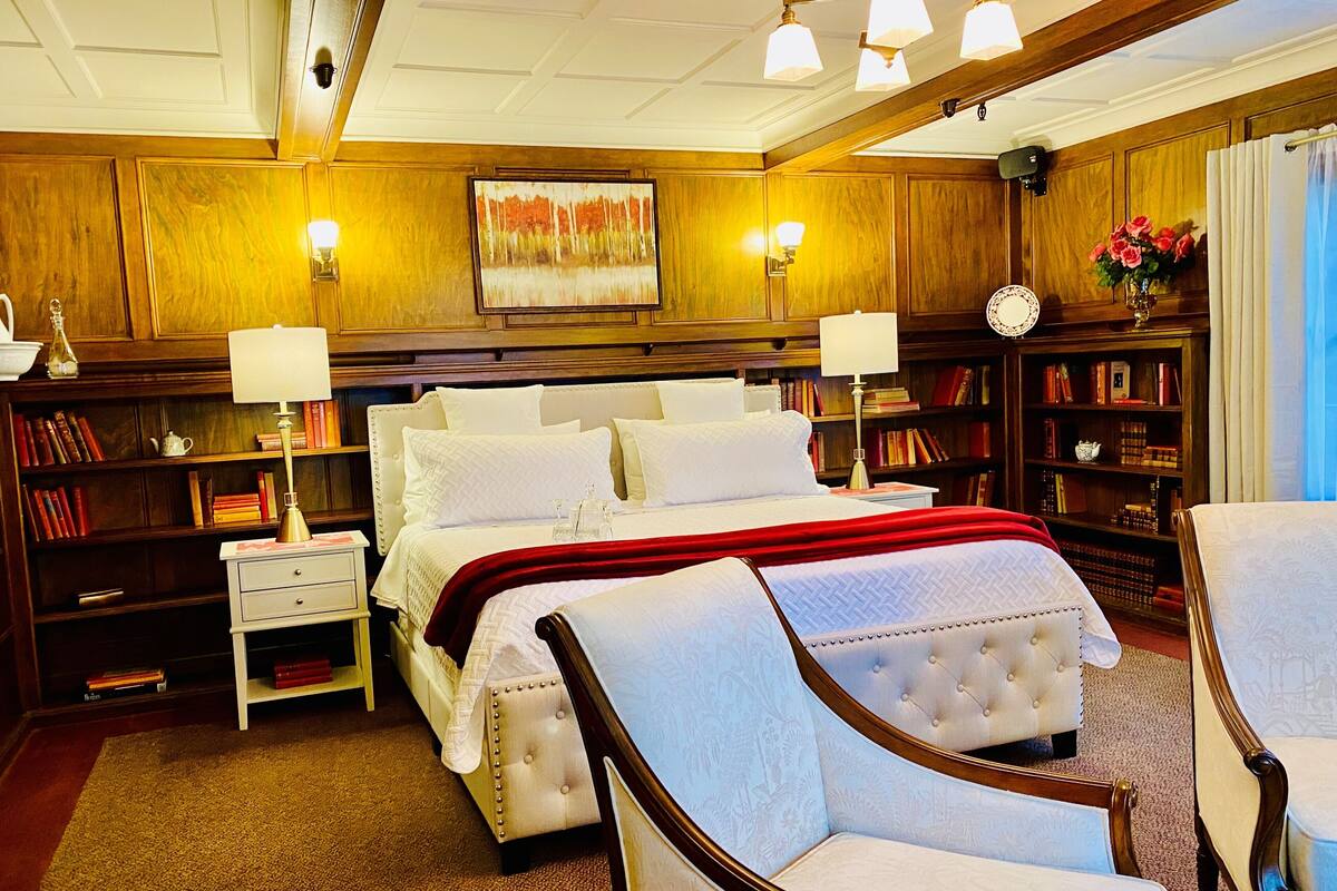 A spacious bedroom with a large bed, bedside tables, a reading chair, and a bookshelf filled with books.