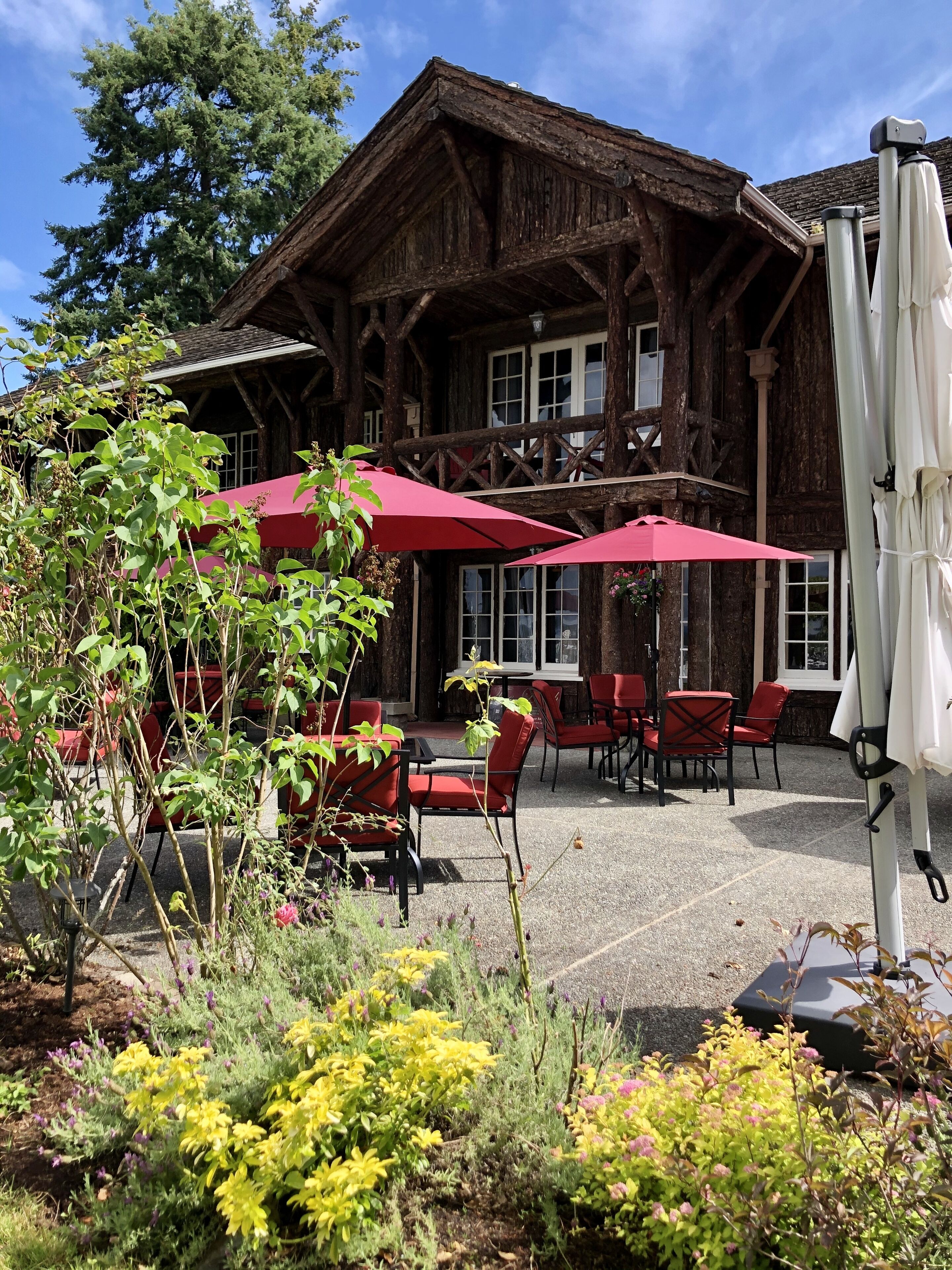 A rustic wooden building with a balcony, outdoor seating with red chairs and umbrellas, and a garden with various plants.