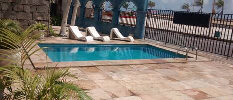 A swimming pool with white lounge chairs, a stone wall, and a metal fence.