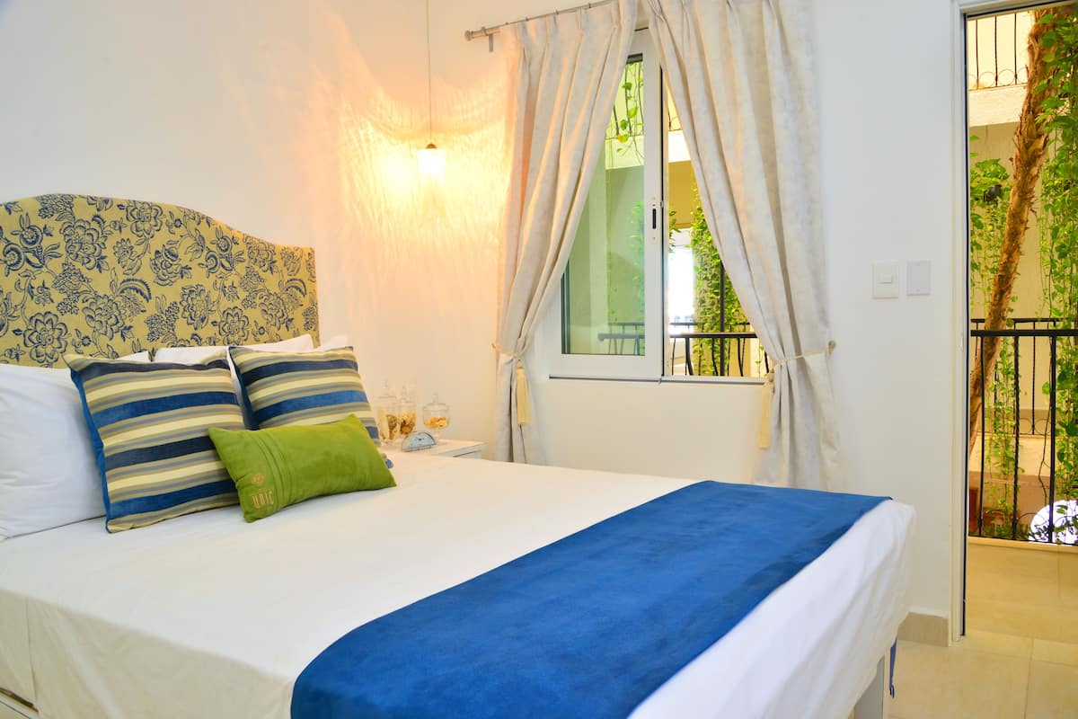 A neatly made bed with a blue bedspread, striped pillows, and a green accent pillow. There is a window with curtains, a bedside table with a lamp, and a view of greenery outside.