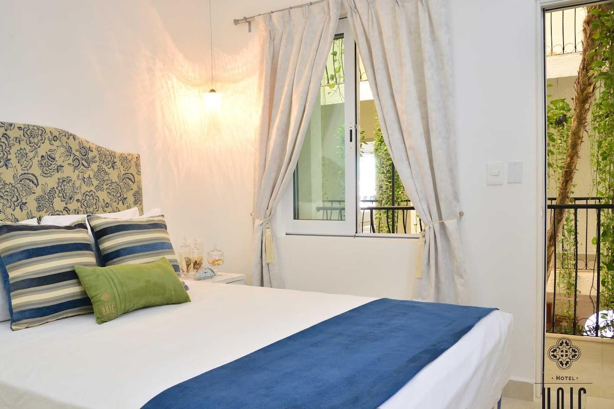A bed with a blue bedspread, striped pillows, and a green pillow. There is a window with curtains, a bedside table with a lamp, and a view of greenery outside.