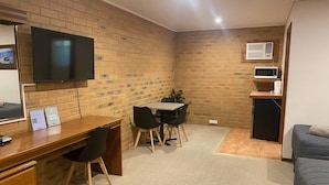 A compact room with a brick wall, a wooden desk with a chair, a small table with chairs, a television mounted on the wall, a microwave, and a refrigerator. - Quays Motel San Remo (San Remo)