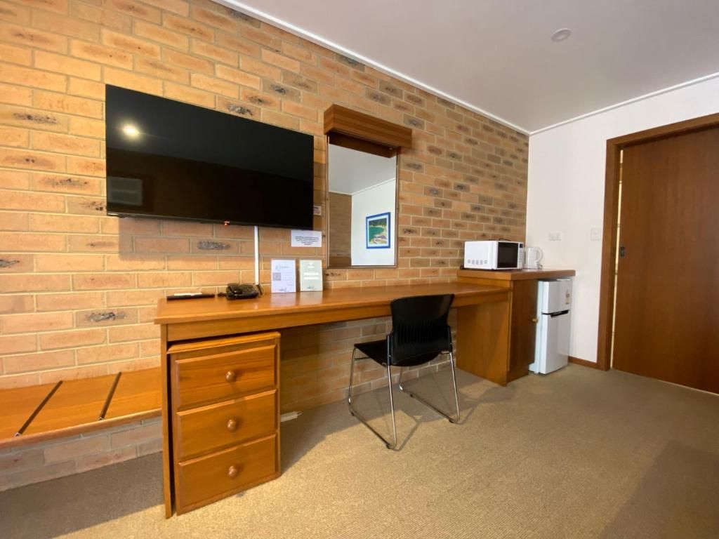 A room with a brick wall, a flat-screen TV mounted above a wooden desk, a black chair, and a microwave.