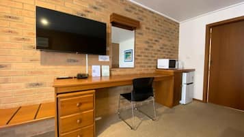 A room with a brick wall, a flat-screen TV mounted above a wooden desk, a black chair, and a microwave.
