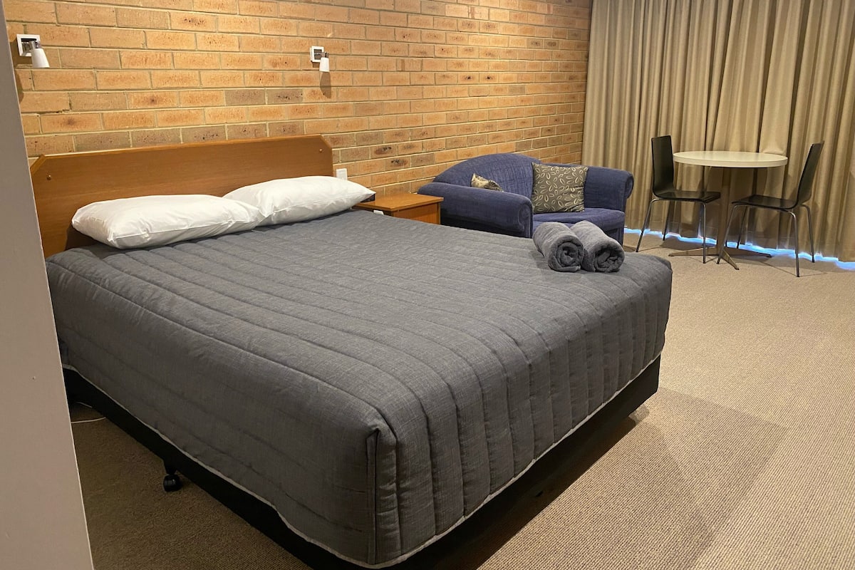 Une chambre d’hôtel avec un grand lit, une tête de lit en bois, un canapé bleu, une petite table ronde et un rideau.