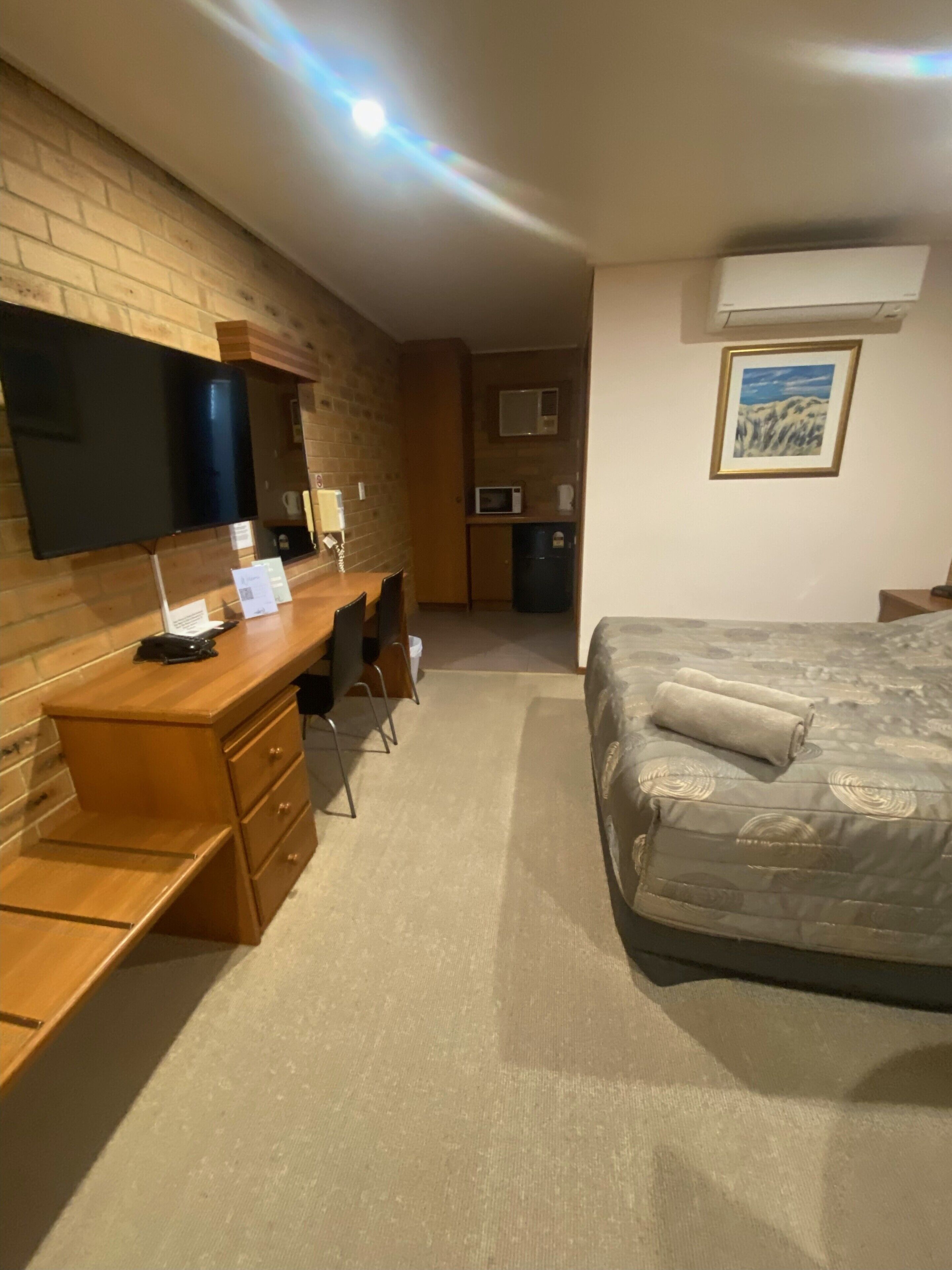 Ein Hotelzimmer mit Holzschreibtisch, einem Bett, einem an der Wand befestigten Fernseher und einer kleinen Küchenzeile.