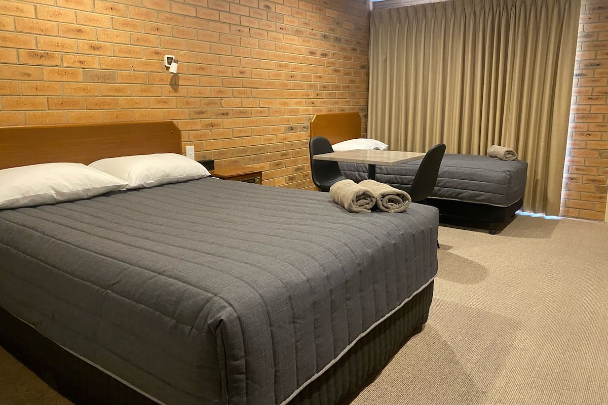 Une chambre d’hôtel avec un mur en briques, un grand lit recouvert d’une couette grise, un plus petit lit, une table de chevet en bois, une table avec une chaise et une fenêtre avec des rideaux.
