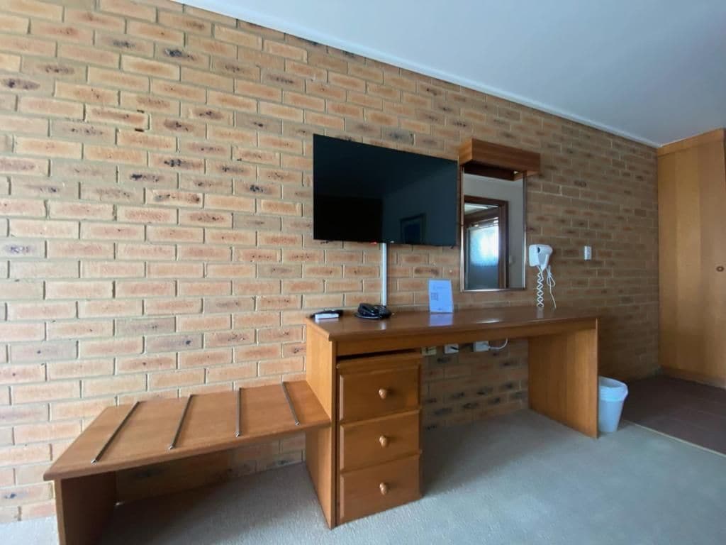 A room with a brick wall, a wooden desk with drawers, a flat-screen TV mounted above it, and a mirror.