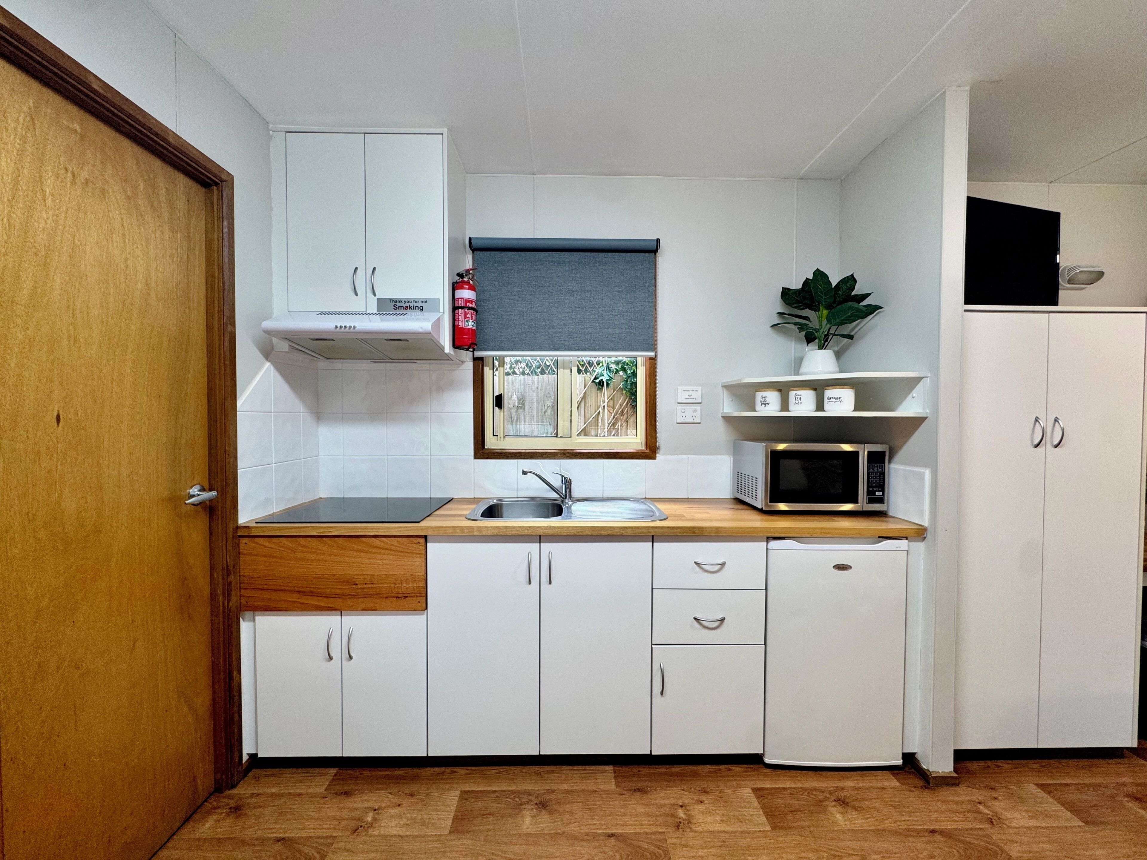 Cocina compacta con armarios blancos, encimera de madera, microondas y un ventanal con vistas a la vegetación.