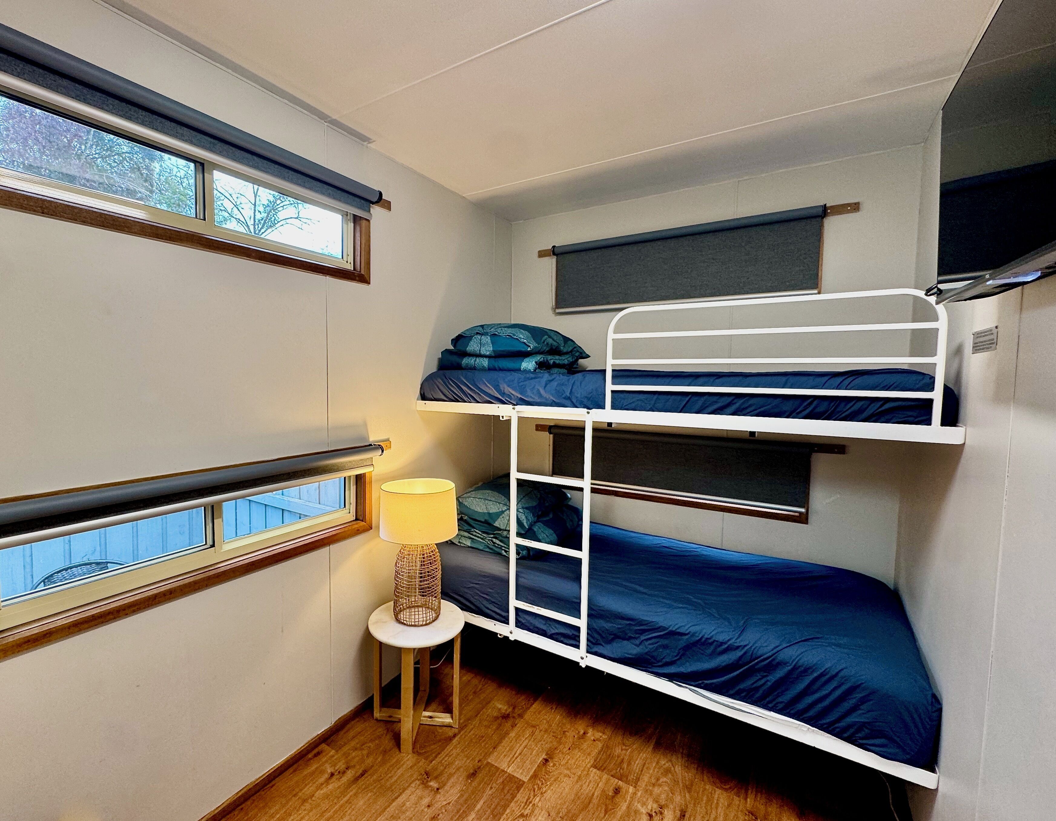 Ett rum med en våningssäng, ett litet bord och en lampa, trägolv och fönster med persienner.