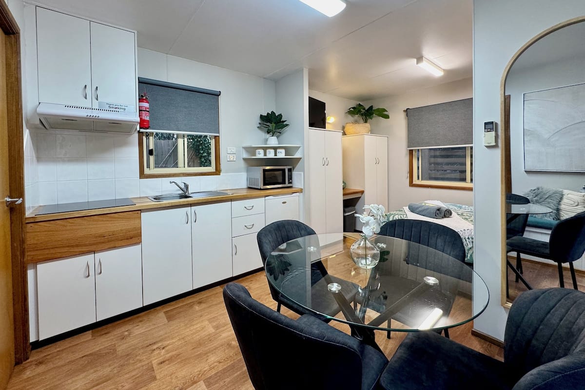 A compact kitchen with white cabinets, a microwave, and a sink. There is a dining area with a glass table and black chairs.