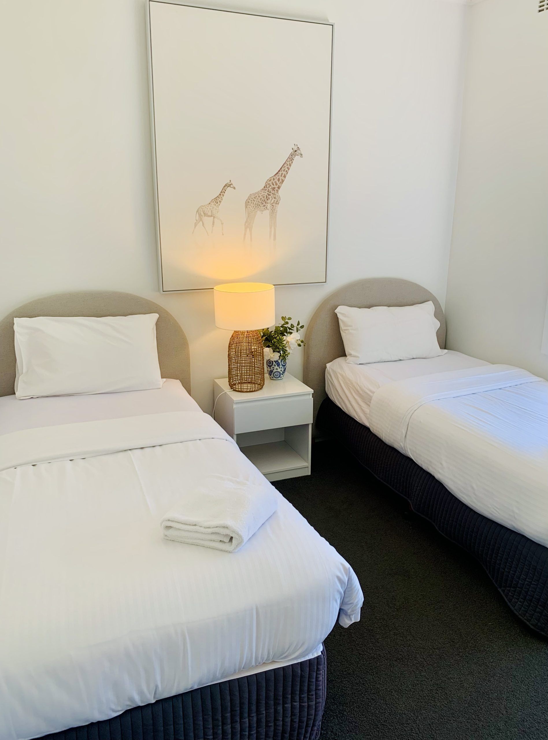 A hotel room with two beds, white linens, a small bedside table with a lamp, and a wall-mounted giraffe artwork.