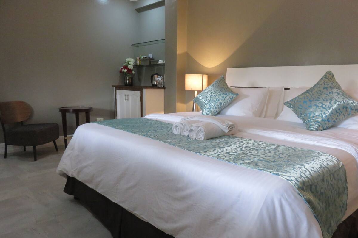 A hotel room with a large bed, a chair, a small table, a cabinet, and a mirror.