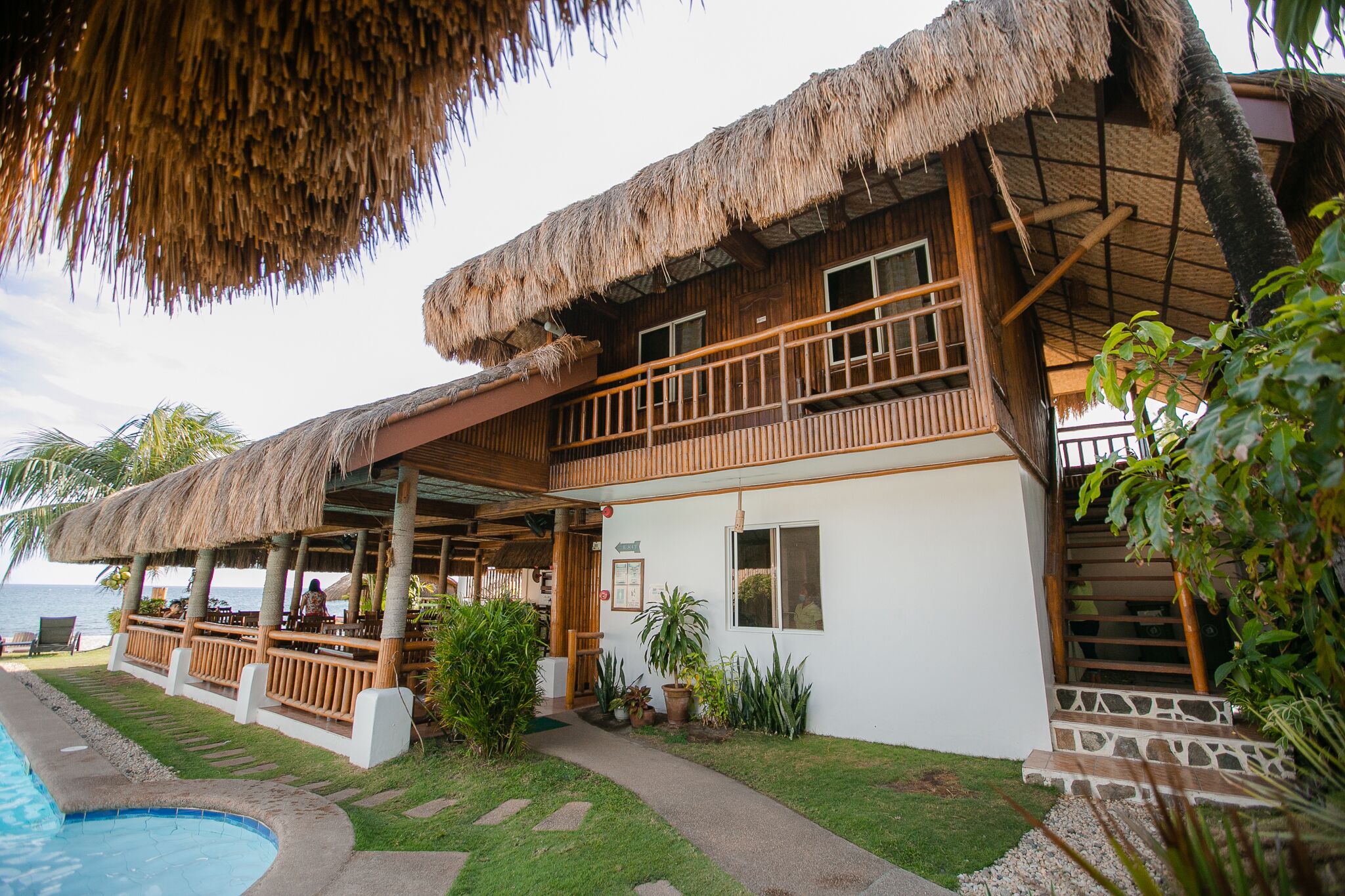 A two-story resort with a thatched roof, a pool, and a view of the ocean.