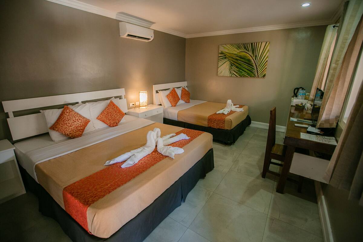 A hotel room with two beds, each with a white headboard, orange patterned pillows, and a red bedspread. There is a desk with a chair, a small table, and a mirror.