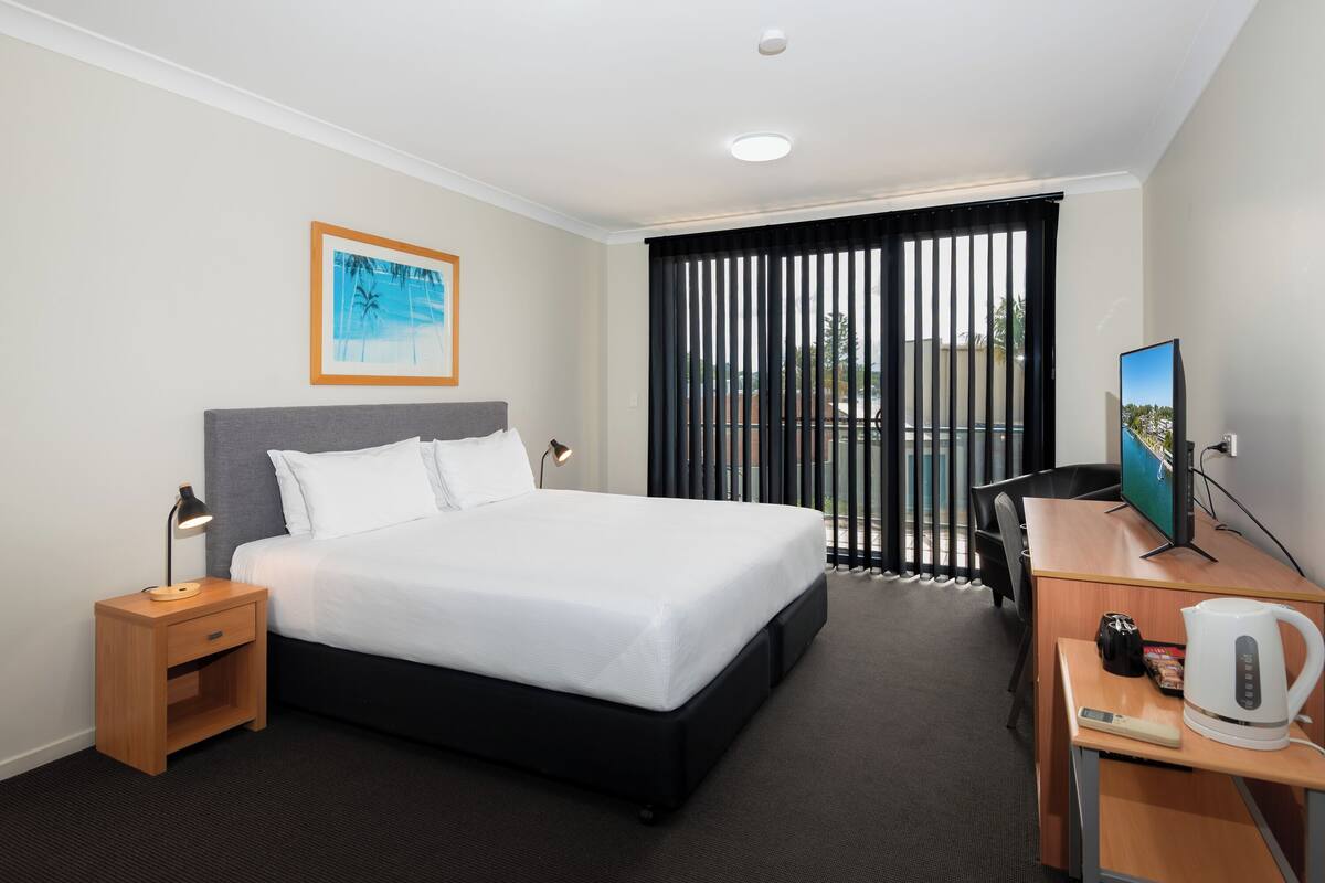A hotel room with a bed, a desk with a computer monitor, a coffee maker, and a picture of a beach.