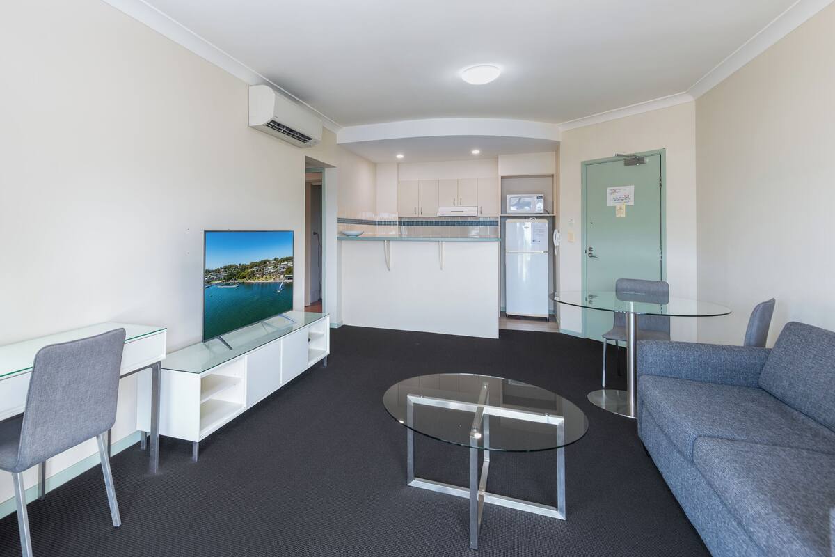 A compact living space with a kitchenette, a glass dining table, a sofa, and a TV on a stand.
