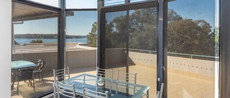 A modern balcony with a glass table and chairs, offering a view of a body of water and distant landscape.