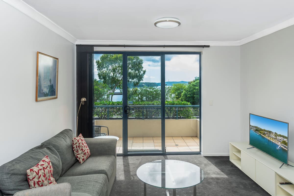 A modern living room with a grey sofa, a glass coffee table, a TV on a white stand, and a balcony with a view of greenery and water.