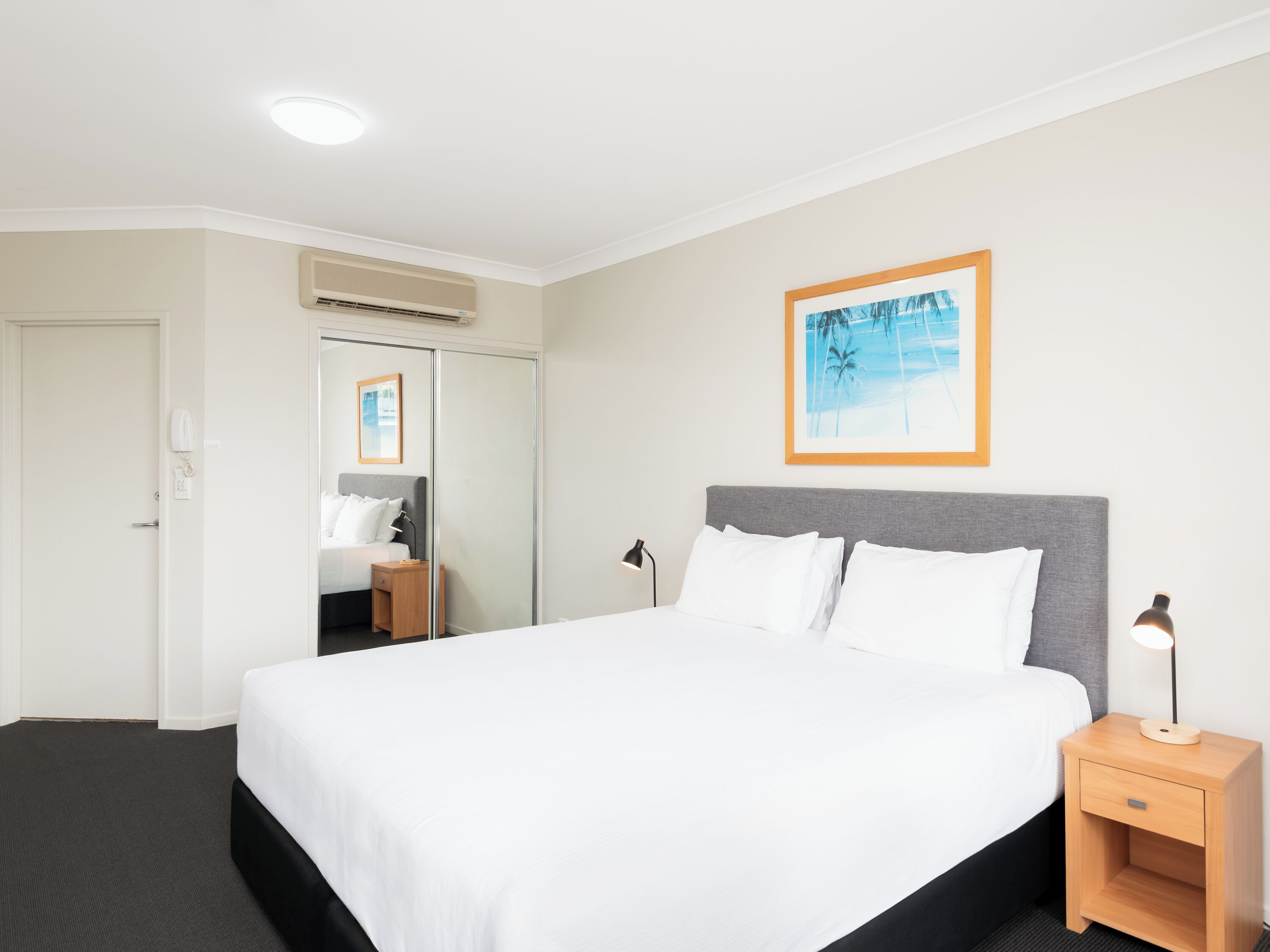Una habitación de hotel con una cama grande, una mesita de noche con lámpara, un cuadro enmarcado en la pared y una puerta con espejo.