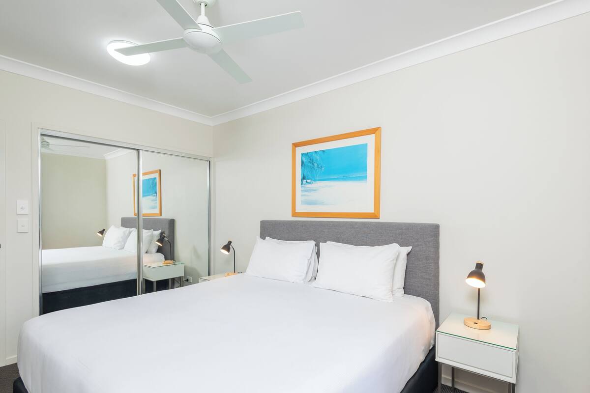 A hotel room with a large bed, a bedside table with a lamp, a ceiling fan, a large mirror, and a framed picture on the wall.