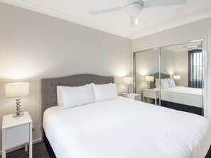 Habitación de hotel ordenada con una cama grande, dos mesitas de noche con lámparas, un ventilador de techo y un espejo grande que refleja la habitación.