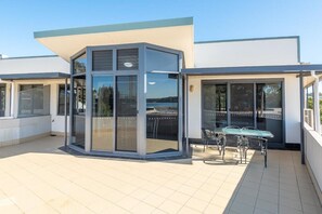 Un edificio moderno con una habitación de esquina con ventanales, un patio con mesa y sillas, y vista al mar.
