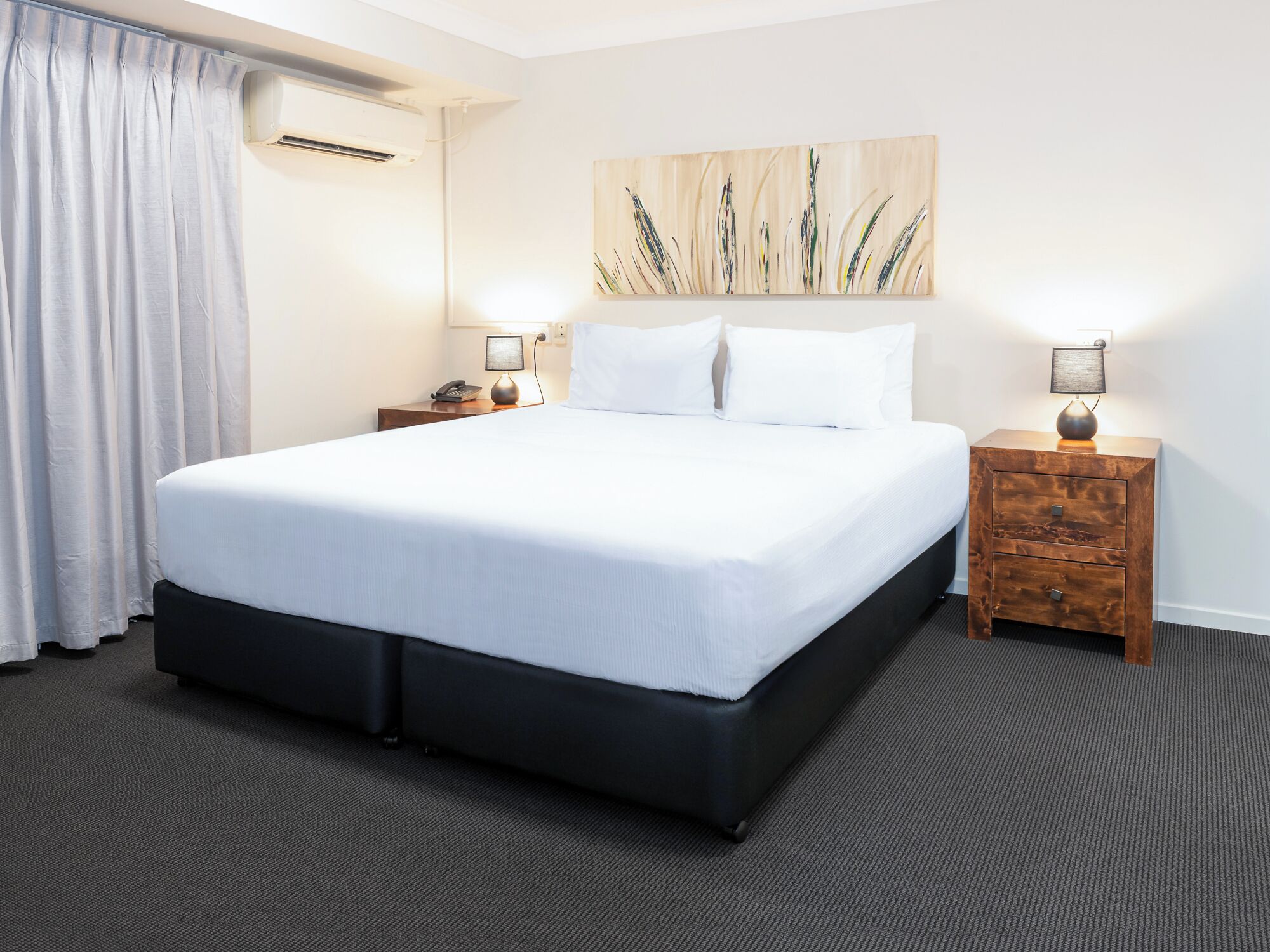 A hotel room with a large bed, two bedside tables with lamps, a painting on the wall, and an air conditioning unit.