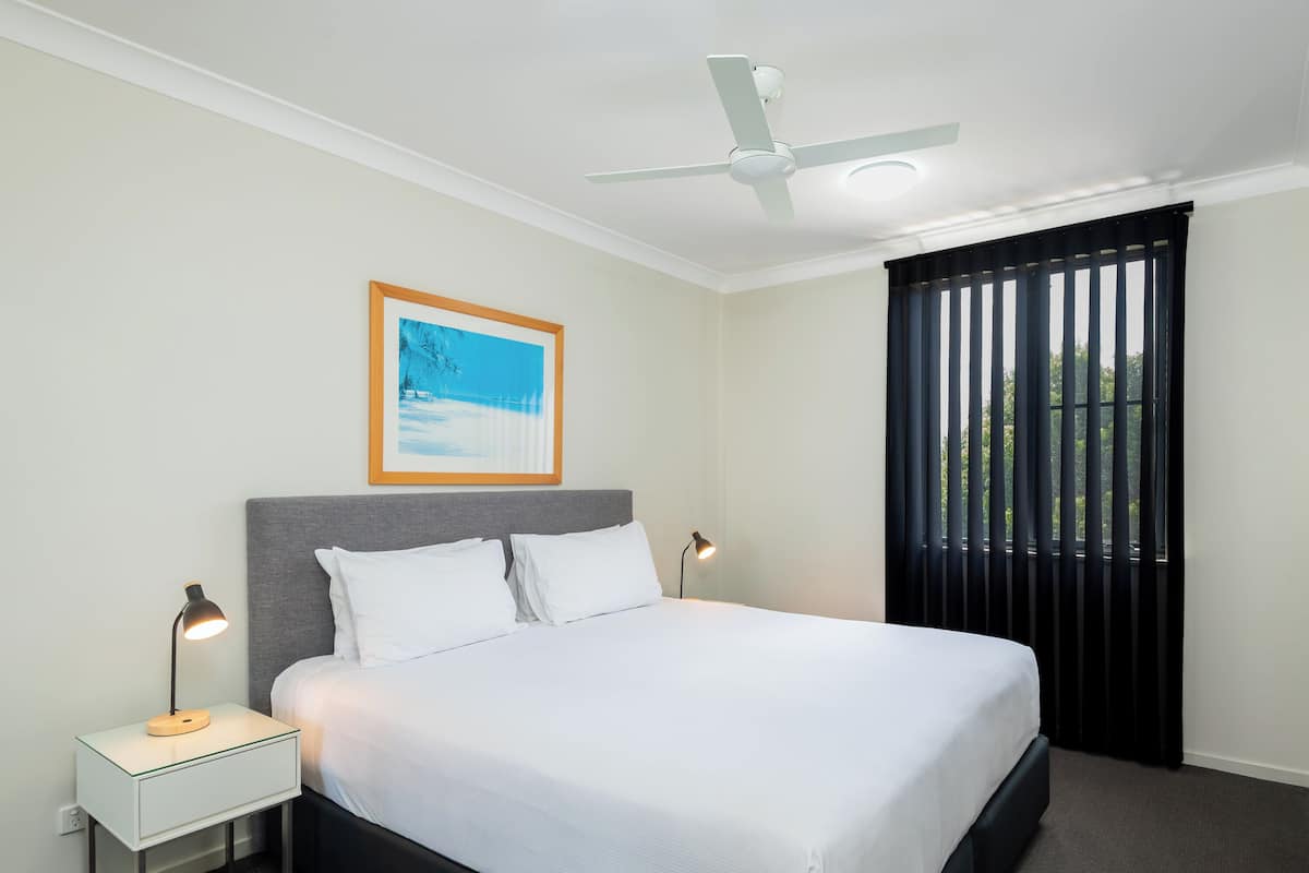 A hotel room with a bed, bedside lamps, a ceiling fan, a framed picture on the wall, and a window with blinds.